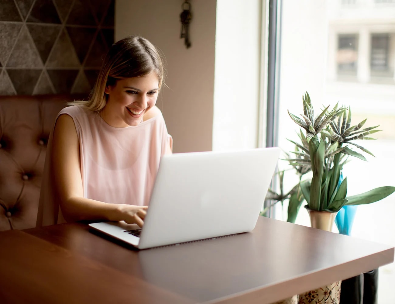 Person smiling while typing on a laptop at a table by a bright window