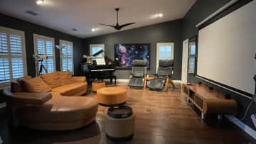 Living area with large projection screen for streaming, baby grand piano that can play on its own, zero gravity chairs to relax your back, and modern sectional sofa.