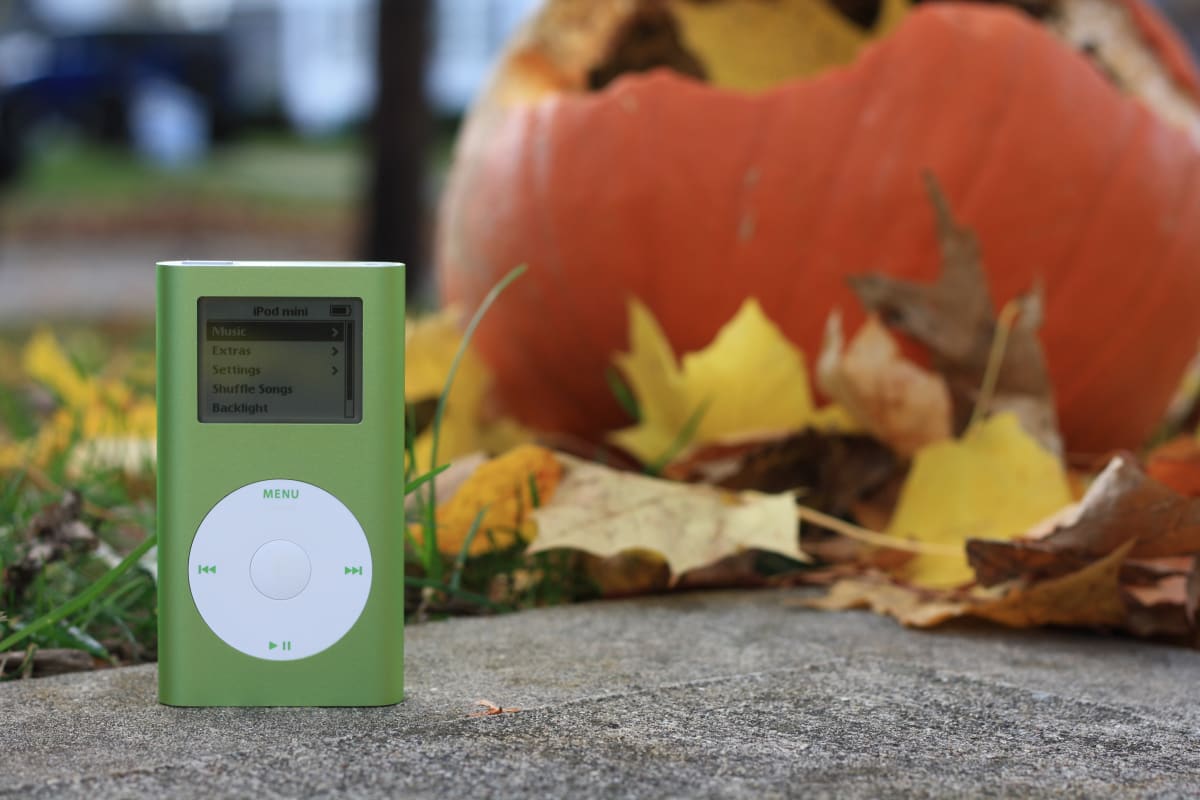 An orange iPod mini sitting on the concrete in front of leaves and a pumpkin in an autumnal scene 