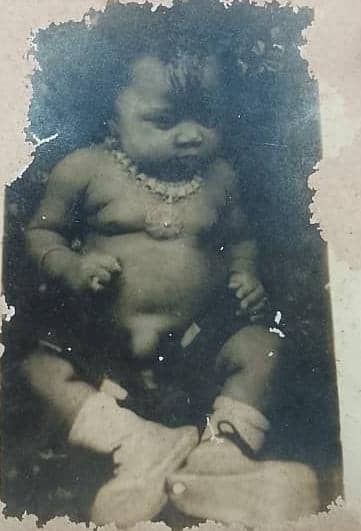 A worn black and white photograph of Oby as a baby, wearing a beaded necklace