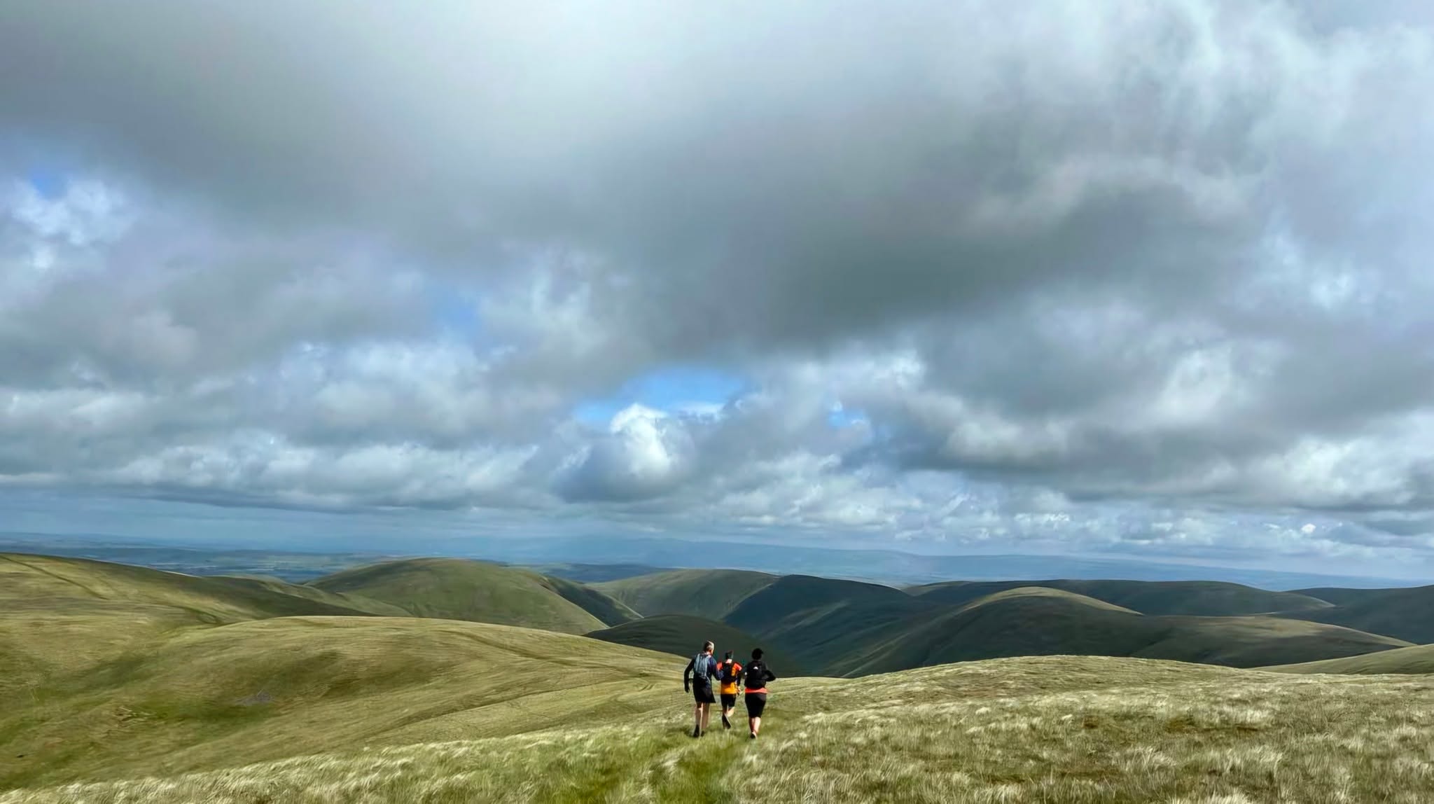 Three UTC runners crossing open green hills beneath a vast sky