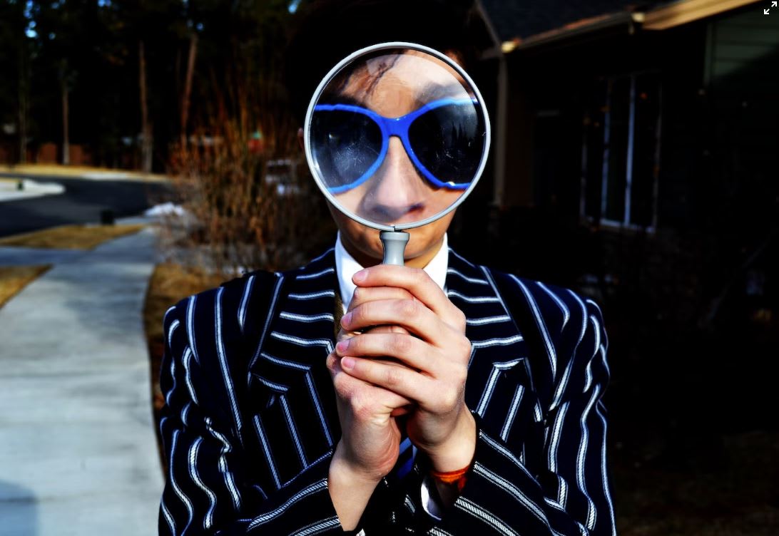 Man in pinstripe suit with blue sunglasses looking through a magnifying glass