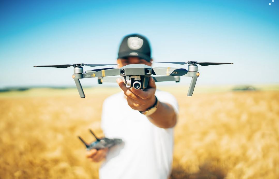 Man in white tshirt and black cap holding drone, against backdrop of a wheat field and clear blue skies