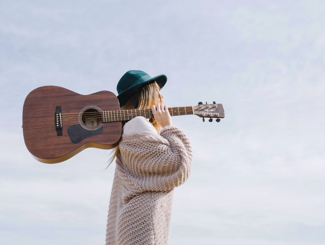 Girl in green hat and chunky knit cardigan holding acoustic guitar over her shoulder