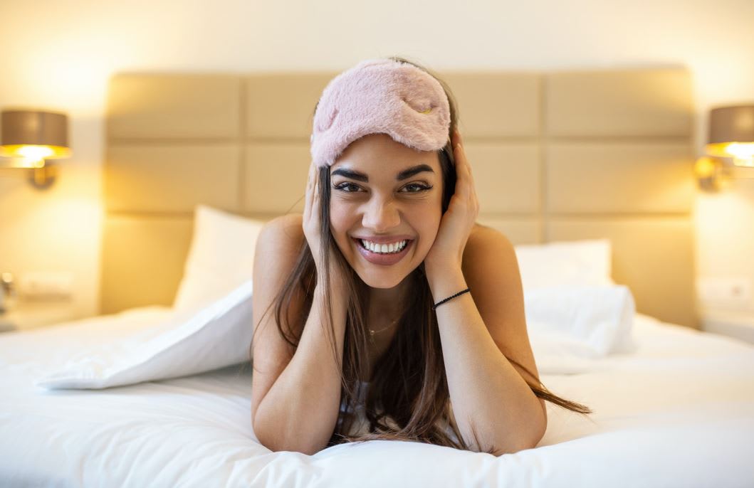 Woman sliming at camera on bed with sleep mask on top of her head