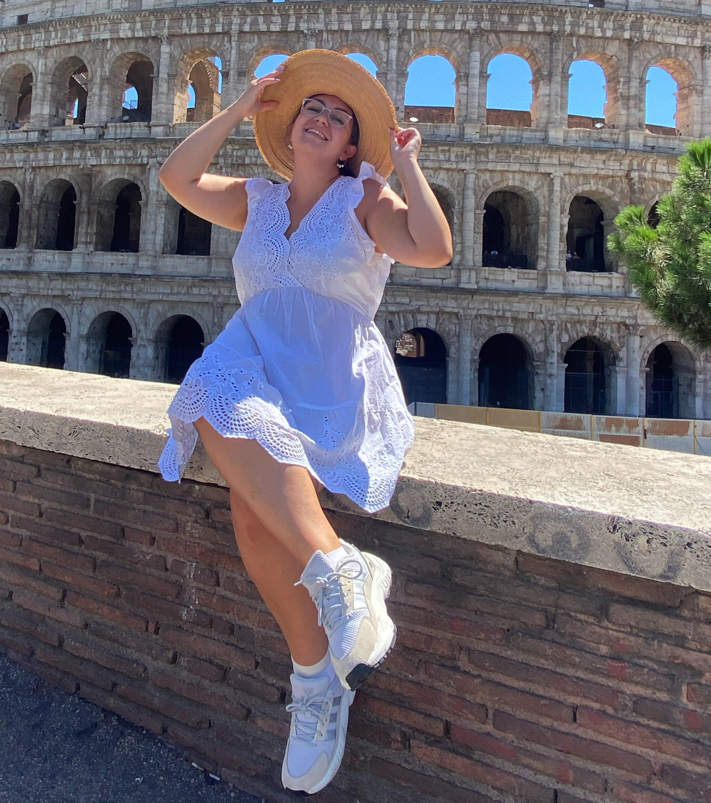 amy lorenz sits on a wall by the Colosseum while wearing a white summer dress and a straw hat
