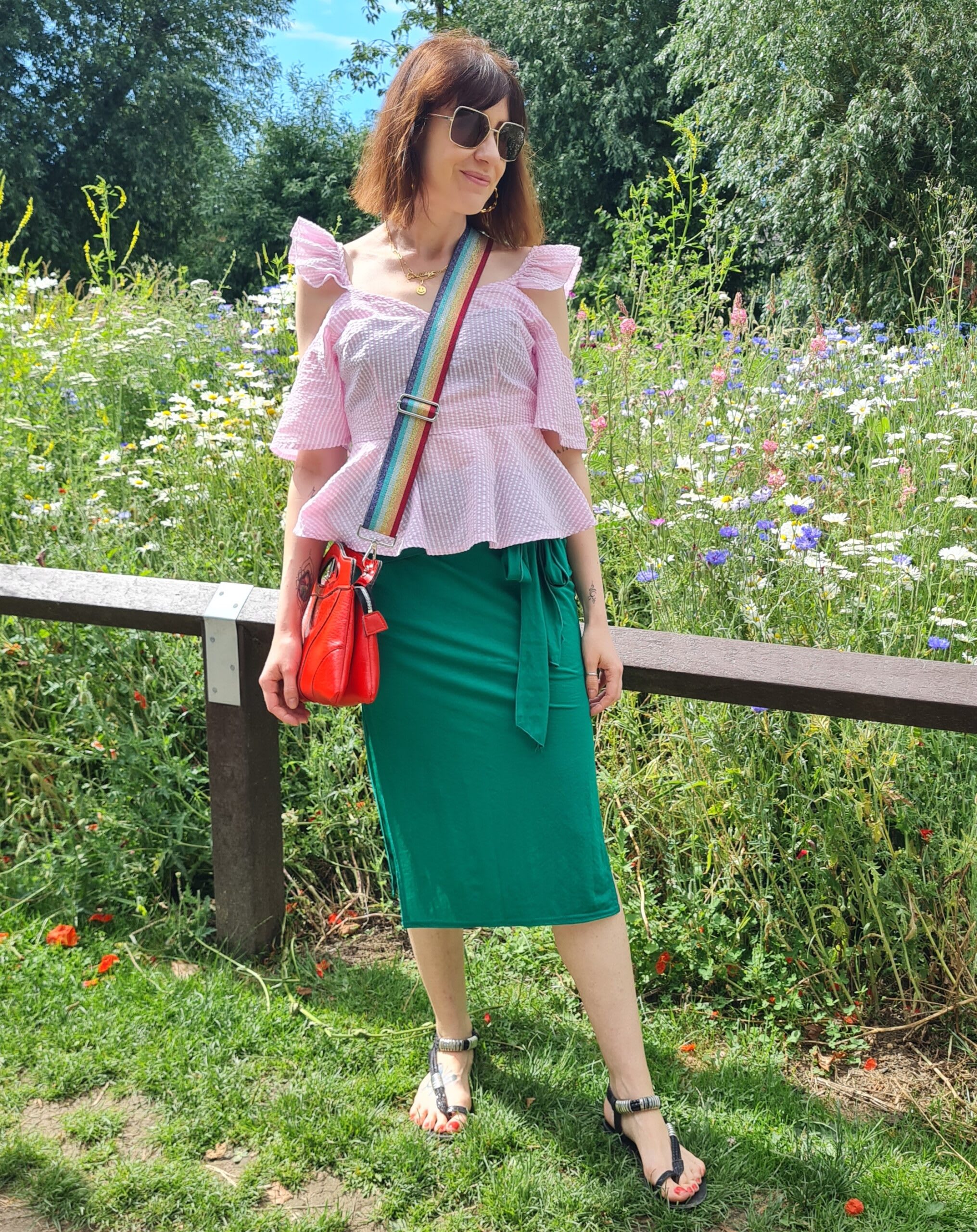 Carola stands by a field of flowers while wearing a pink top with a green pencil skirt