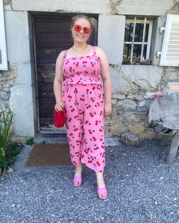 The thrifty edit standing wearing a pink jumpsuit with fruit on it with matching red glasses and bag