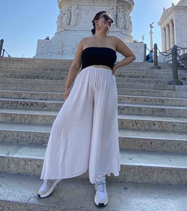 Amy Lorenz standing on some steps while looking up, wearing a wide leg pair of white trousers, a black bandeau top and sunglasses