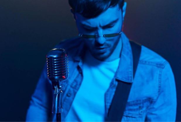 Photo by ANTONI SHKRABA production: https://www.pexels.com/photo/a-man-in-denim-jacket-standing-in-front-of-a-microphone-while-looking-down-8041066/ 