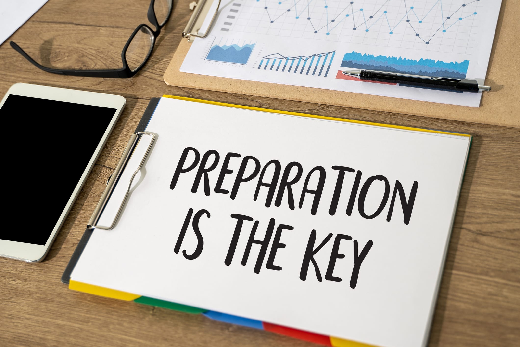 Work desk with a clipboard displaying the message 'Preparation is the key' next to a smartphone, eyeglasses, and graphs on paper, symbolizing strategic planning and organization.