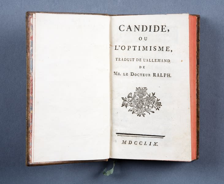 The French 1759 edition of Candide, ou l'Optimisme is open on the title page. It reads “CANDIDE, OR, ALL FOR THE BEST, TRANSLATED FROM GERMAN BY DOCTOR RALF”. Beneath this, there's a printout of an image in which flowers, foliage, a stick and, possibly, a trumpet are all intermingled. At the bottom of the page, there are two horizontal lines, one on top of the other, running parallel to each other, but not quite. The top one is about twice as thick as the bottom one. The width of the two lines is about two-thirds of the page. MDCCLIX is written below these lines. The outer edge of the pages is red, and a coniferous green ribbon-signet protrudes from the book at the bottom center. The book stands vertically on a blue-gray felt-like background.