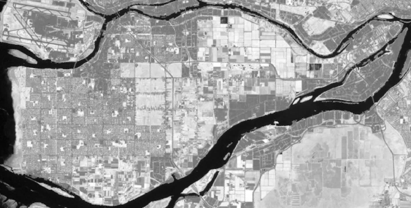 A black-and-white aerial photo of Richmond, British Columbia. The main land mass of the city is narrowly separated from the surrounding area by rivers. The island is heavily urbanized, with distinct roads, residential developments and other land uses visible. Whiter areas are more vegetated whereas darker areas are built-up urban environments.