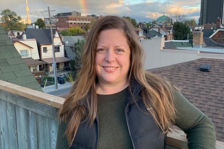 A smiling Caucasian woman with loose, long light brown hair leaning back against a wooden patio rail overlooking a street
