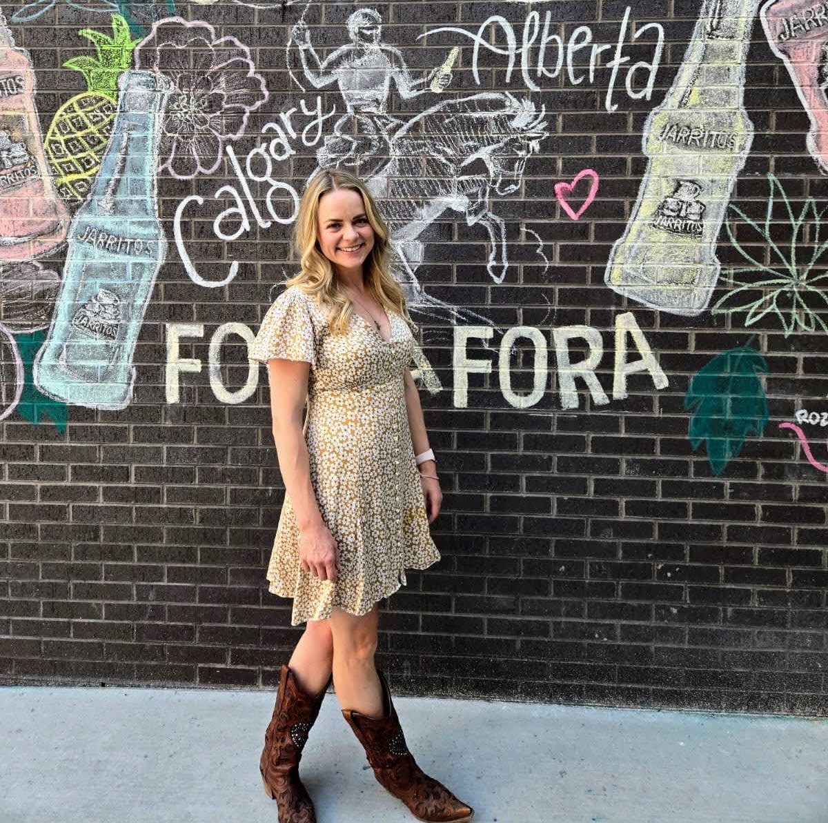 An image showing a blonde woman with shoulder length hair smiling in a dress and cowboy boots.