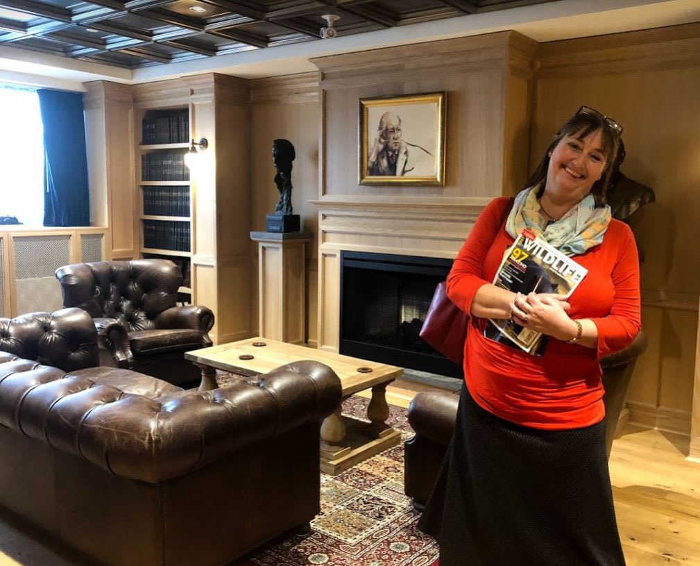 A woman in a red sweater and scarf smiling at the camera in a room with a leather sofa and chair, a fireplace, a book self and painting.