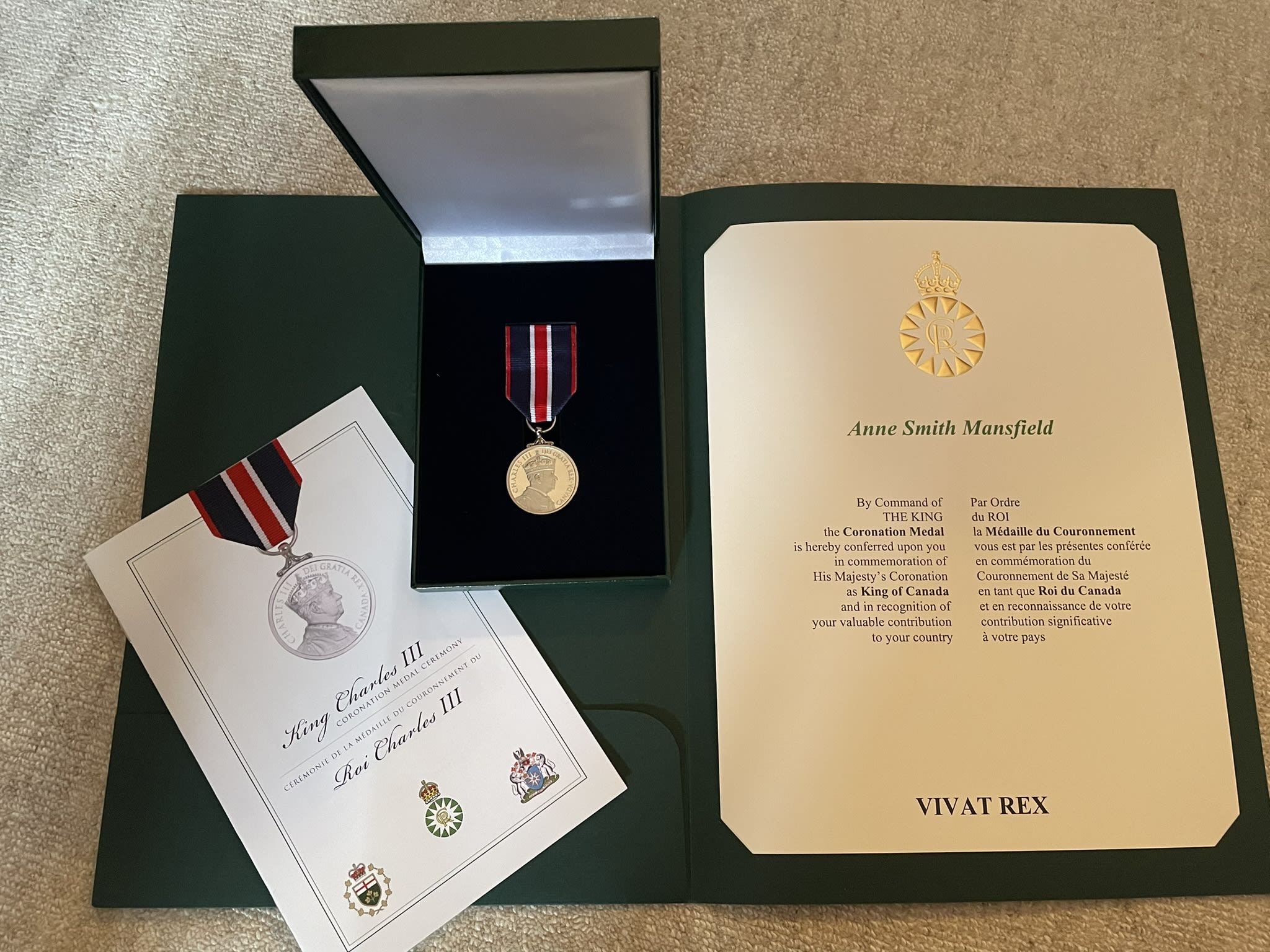 An image of Anne Mansfield’s King Charles III Coronation medal and certificate showing a gold medal and ribbon with King Charles’ profile, and two official documents with her name on it and the official crests of the king and the government of Canada.