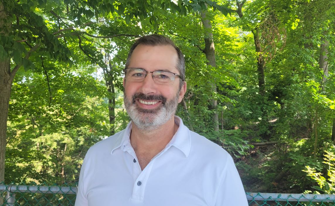 A man with a beard and glasses smiling at the camera in front of a forest.
