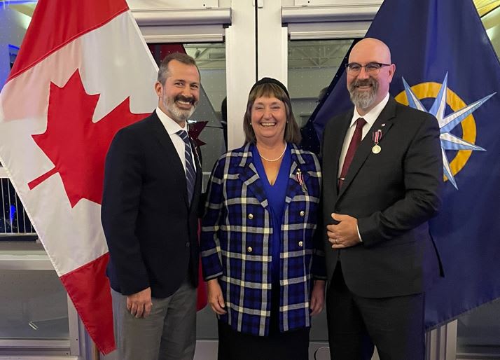 A woman standing between two men in front of a Canadian and a RCGS flag. They are all smiling at the camera.