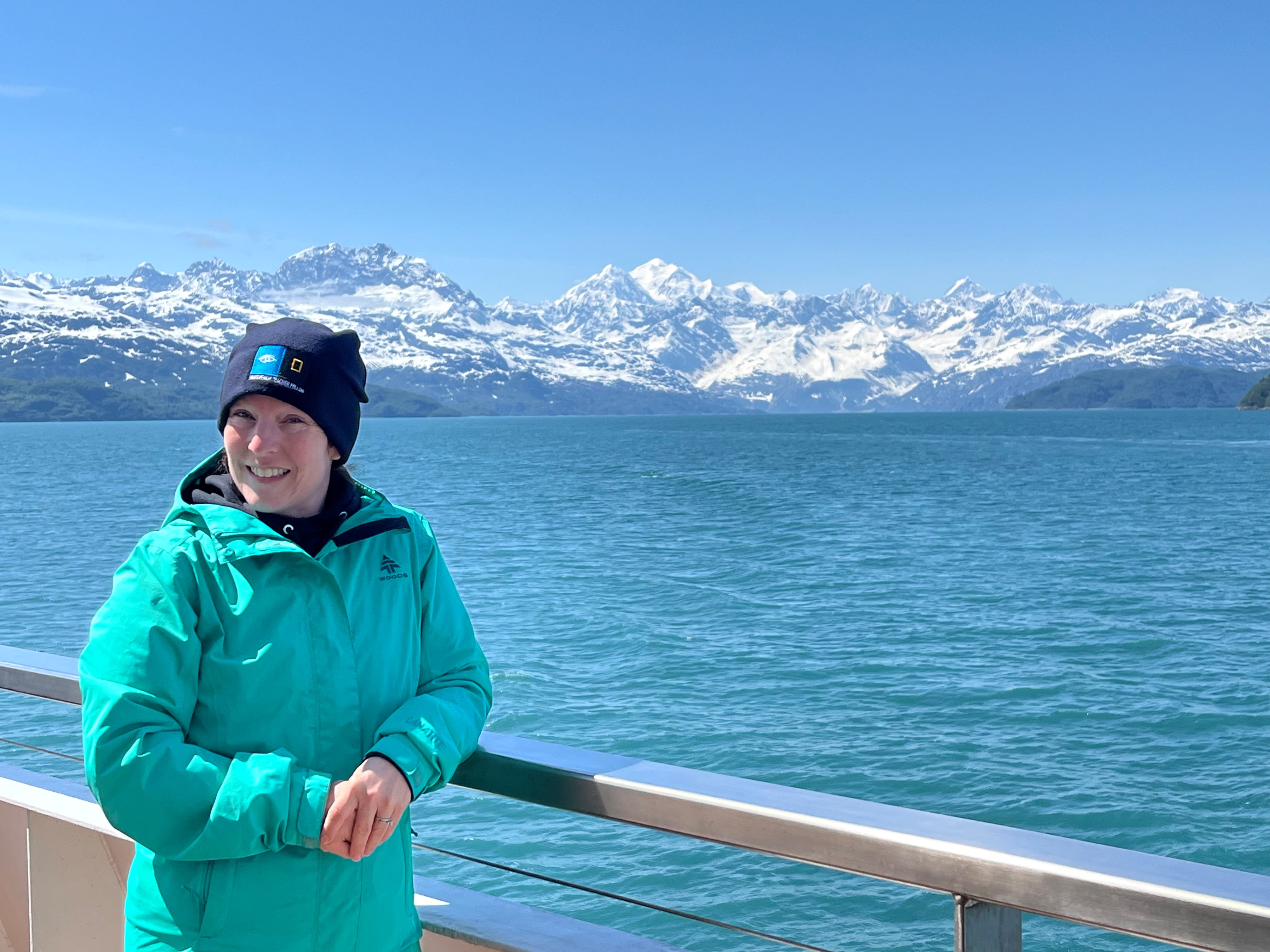 A woman in a toque smiling in front of an ocean and a mountain range.