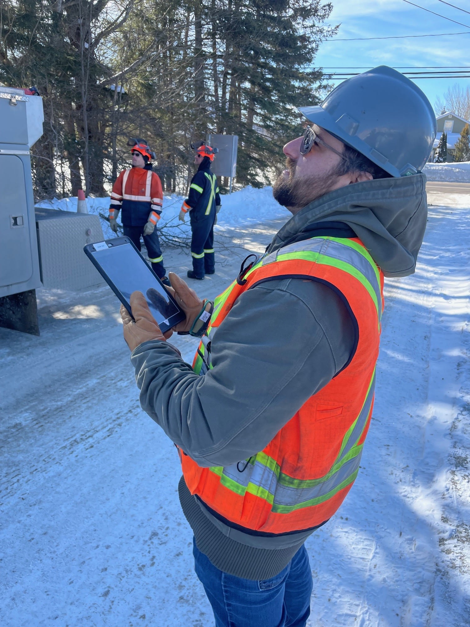 An Edmundston public works crew member stands outside on a snowy winter day, holding a tablet displaying a work order and looking up at the scene.