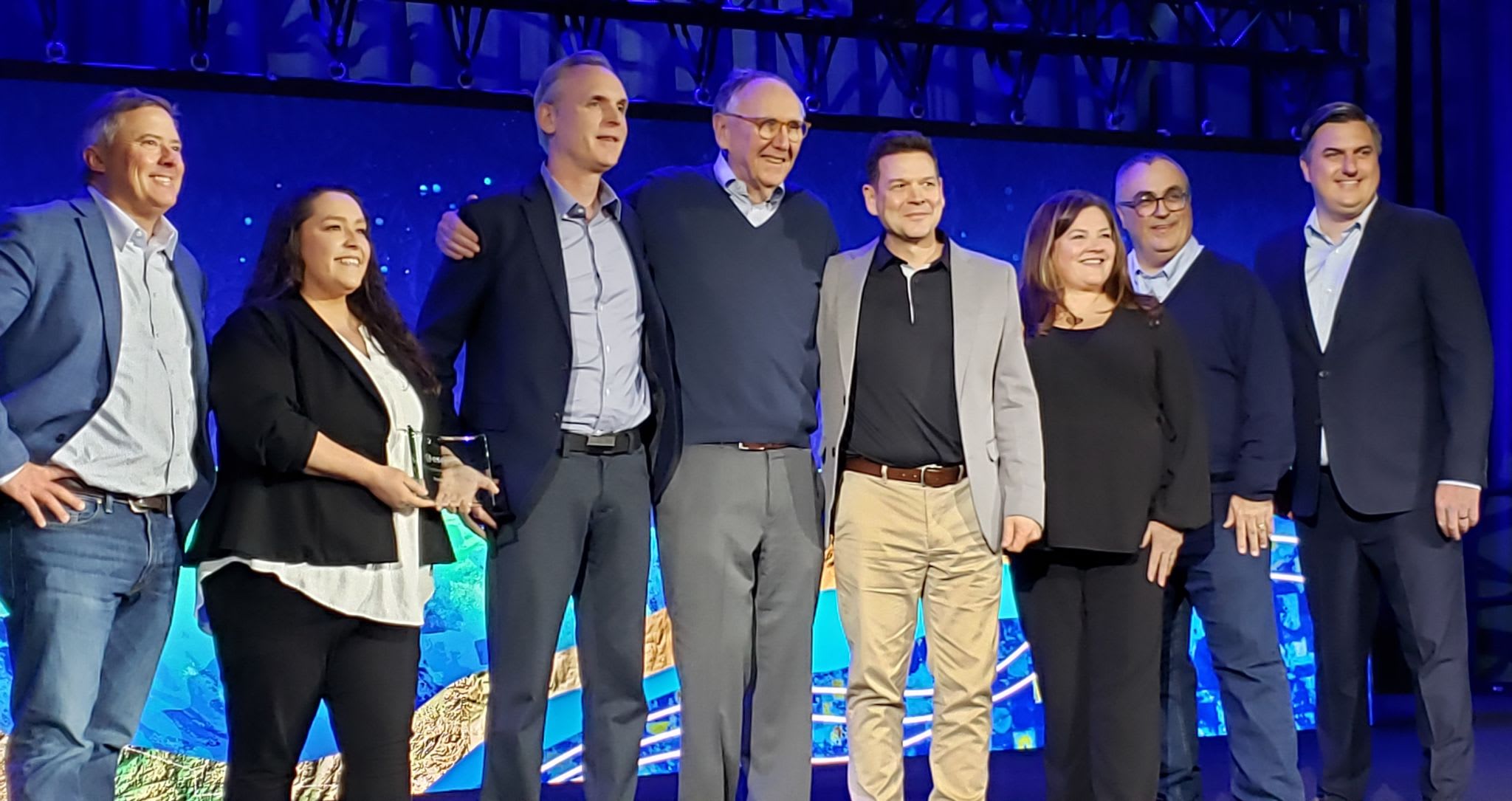 A group of six men and two women smiling on stage. One of the women is holding a glass award plaque.