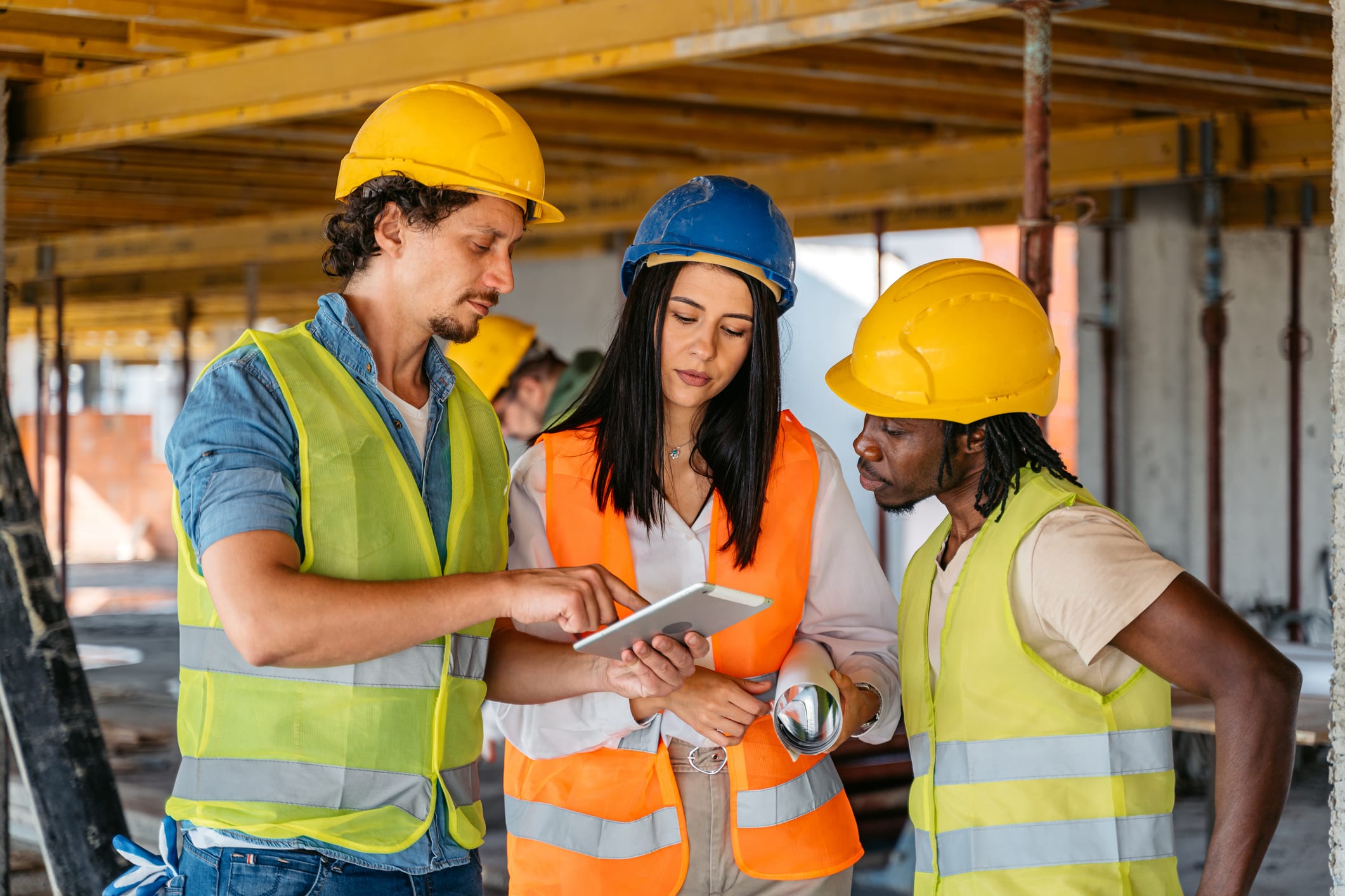 Construction worker, architect and engineer wearing hard hats and safety gear on a worksite, all looking at a tablet.