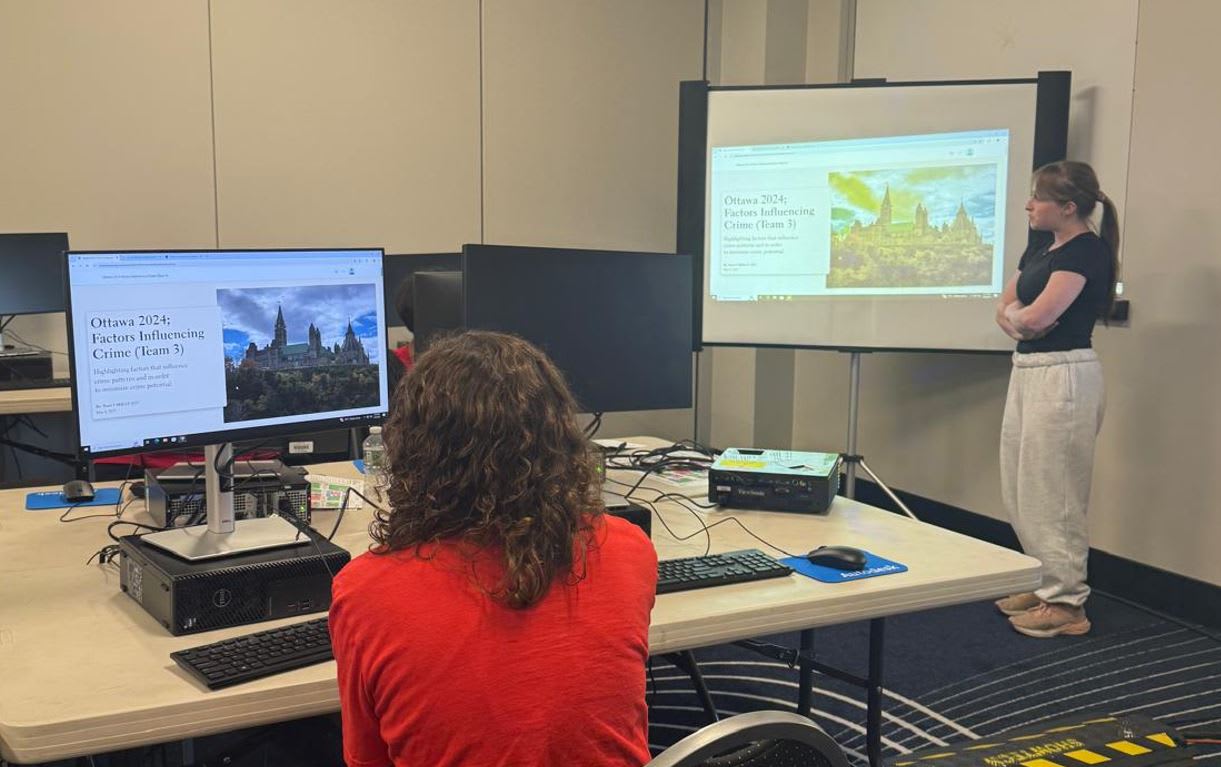 Image shows two female students presenting their work. One student is sitting at a computer, and the other student is standing speaking to the judges.