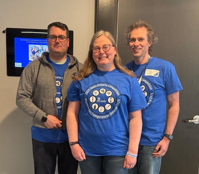 An image showing three people. Two men and a woman standing in front of a screen.