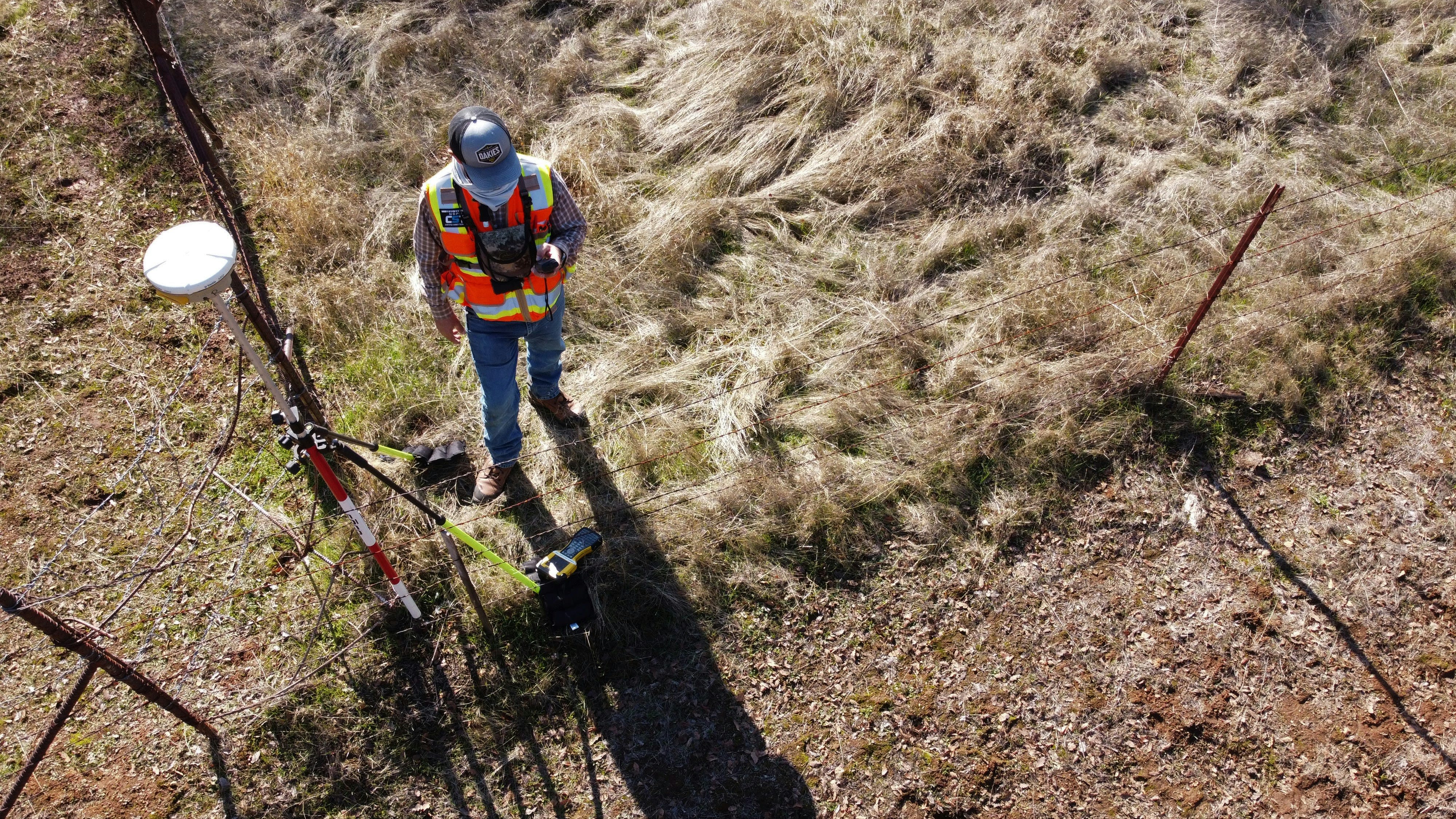 Civil engineer in safety gear using GNSS surveying equipment near a wire fence to collect geospatial data in a grassy field.