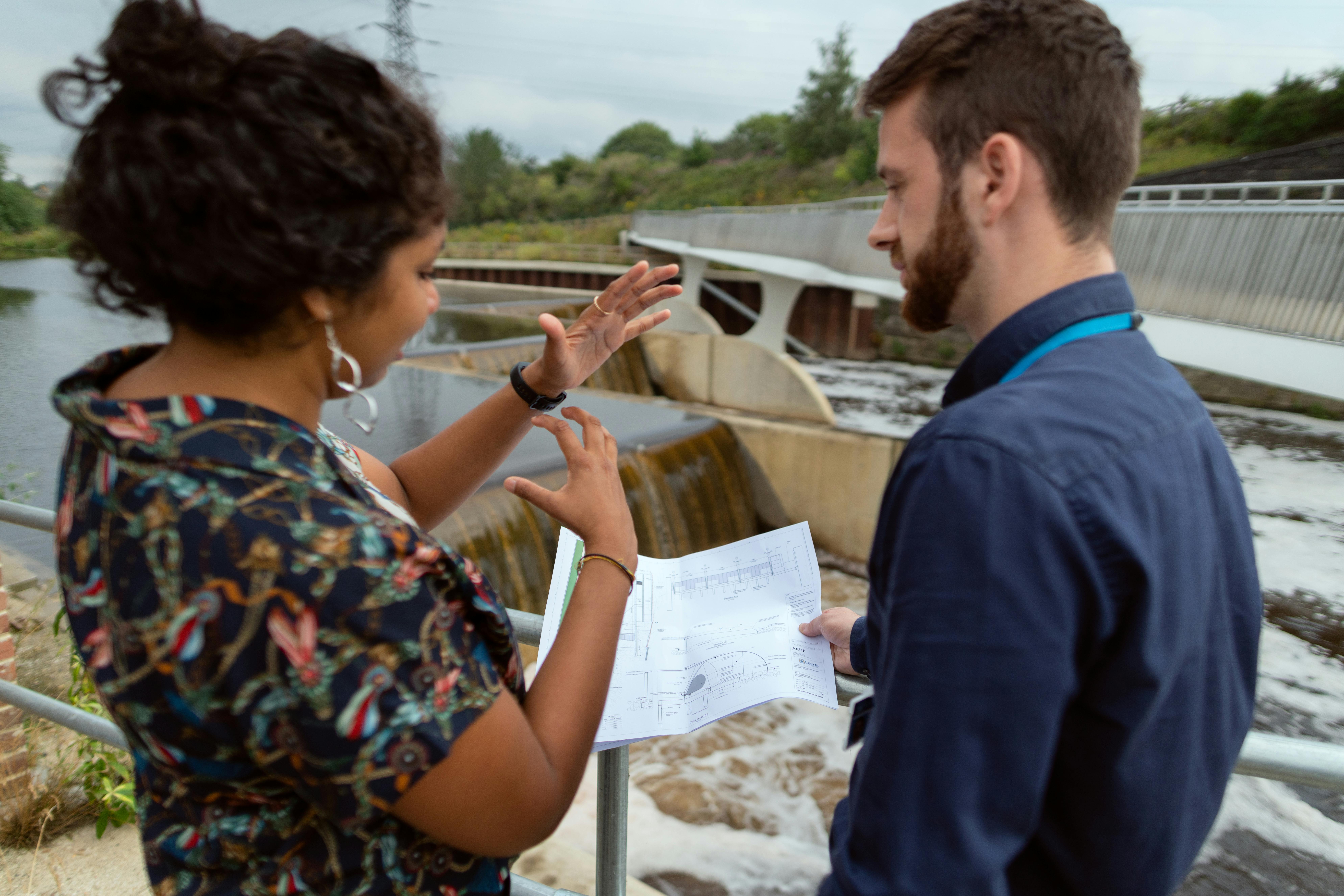Two engineers reviewing documents while inspecting water treatment plant infrastructure, with treatment tanks and walkways visible in the background.