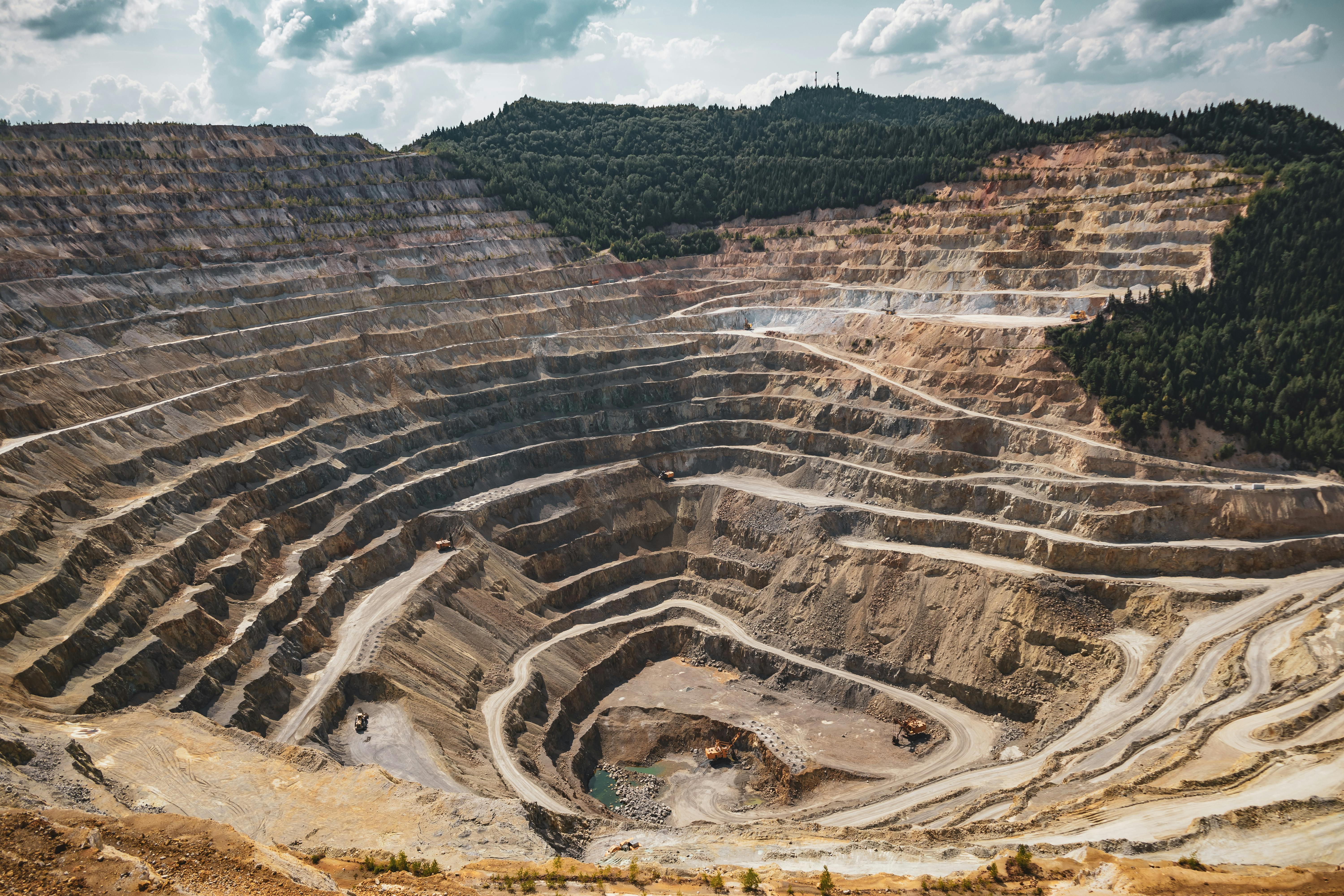 Wide view of a terraced open-pit mine with haul trucks on multiple levels, surrounded by forested hills and a partly cloudy sky.