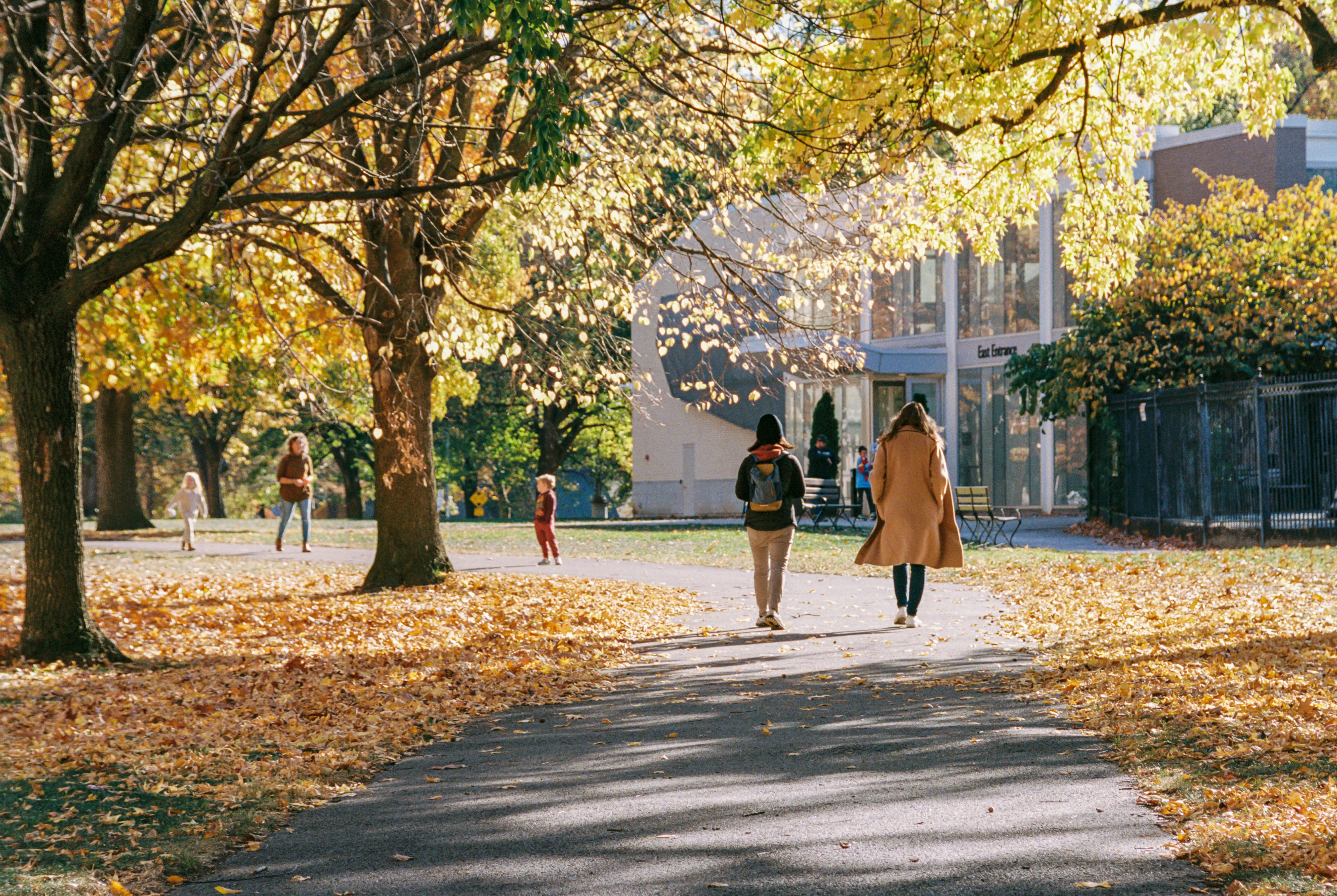 A fall scene with leaves on the ground and people walking towards a building.