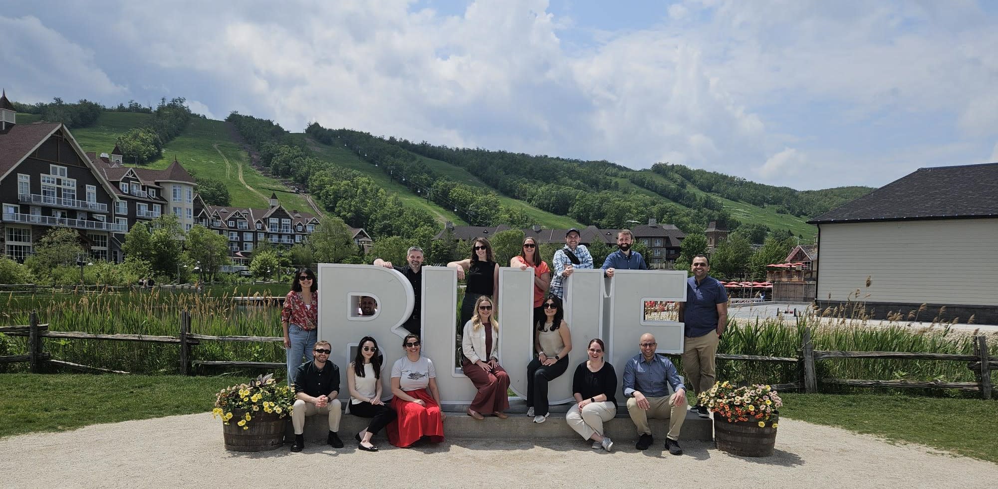 Photo de groupe de l’équipe Milieu scolaire et recherche à Inspire 2025. Les membres de l’équipe sont debout et assis autour du grand monument « BLUE » à Blue Mountain. À l’arrière-plan, on aperçoit des collines verdoyantes et des bâtiments sous un ciel nuageux.