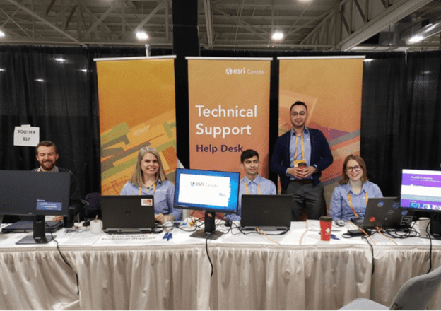 The Technical Support Help Desk at an Esri Canada User Conference. Four Esri Canada team members in matching pale blue button-down shirts sit smiling at a long table with a white tablecloth and computer equipment on it. Behind them are three orange vertical banners against a dark curtained backdrop, as well as one more member of the team who is standing and smiling at the camera. 