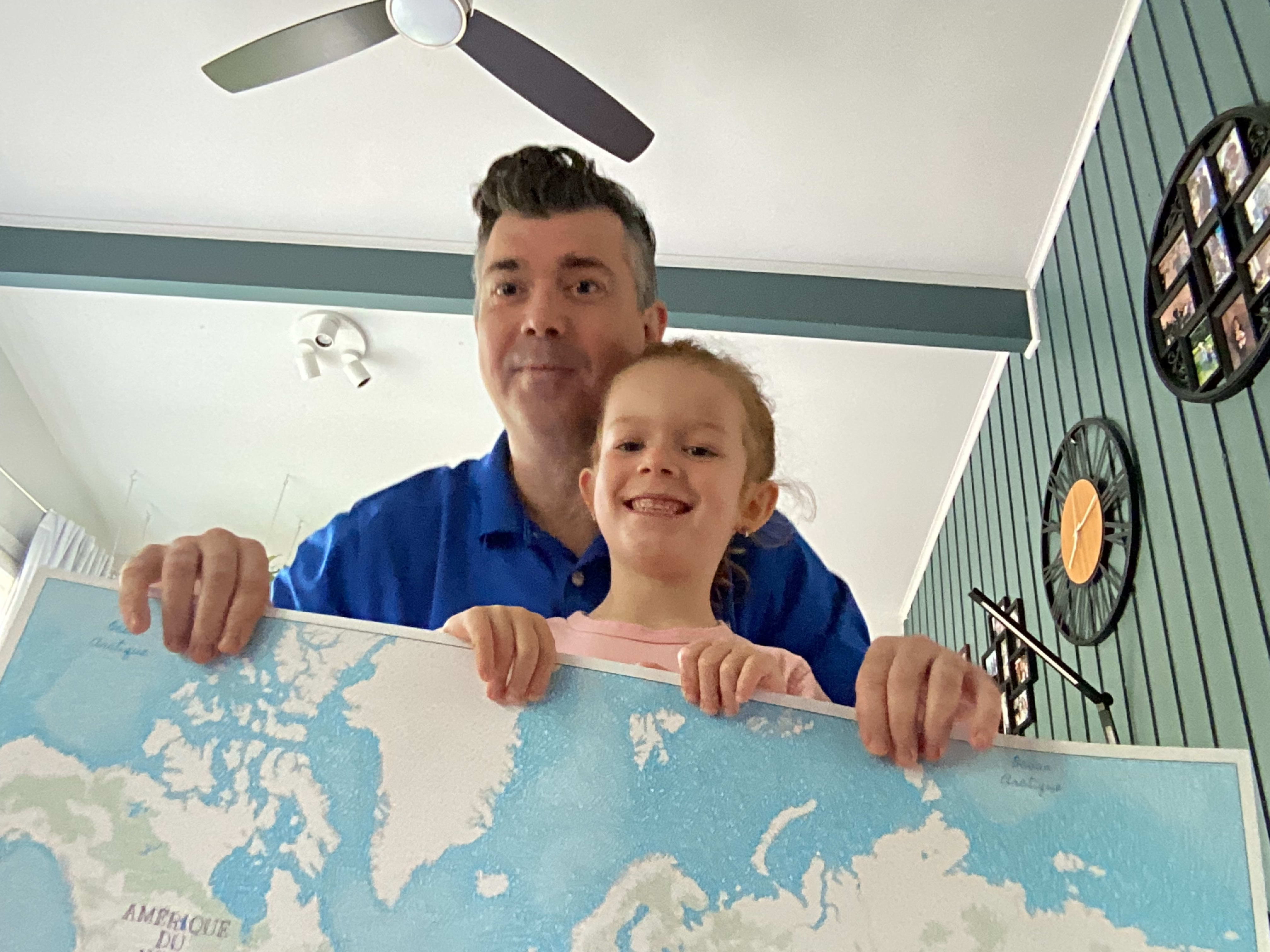 Albert Chifoi and his daughter are holding a printed world map that was used during a classroom activity. Only about one-third of the map is visible in the image; the rest is out of frame. The northern part of North America, Greenland, Iceland, several northern European countries and part of northern Russia can be seen. The map is in a hand-drawn style.