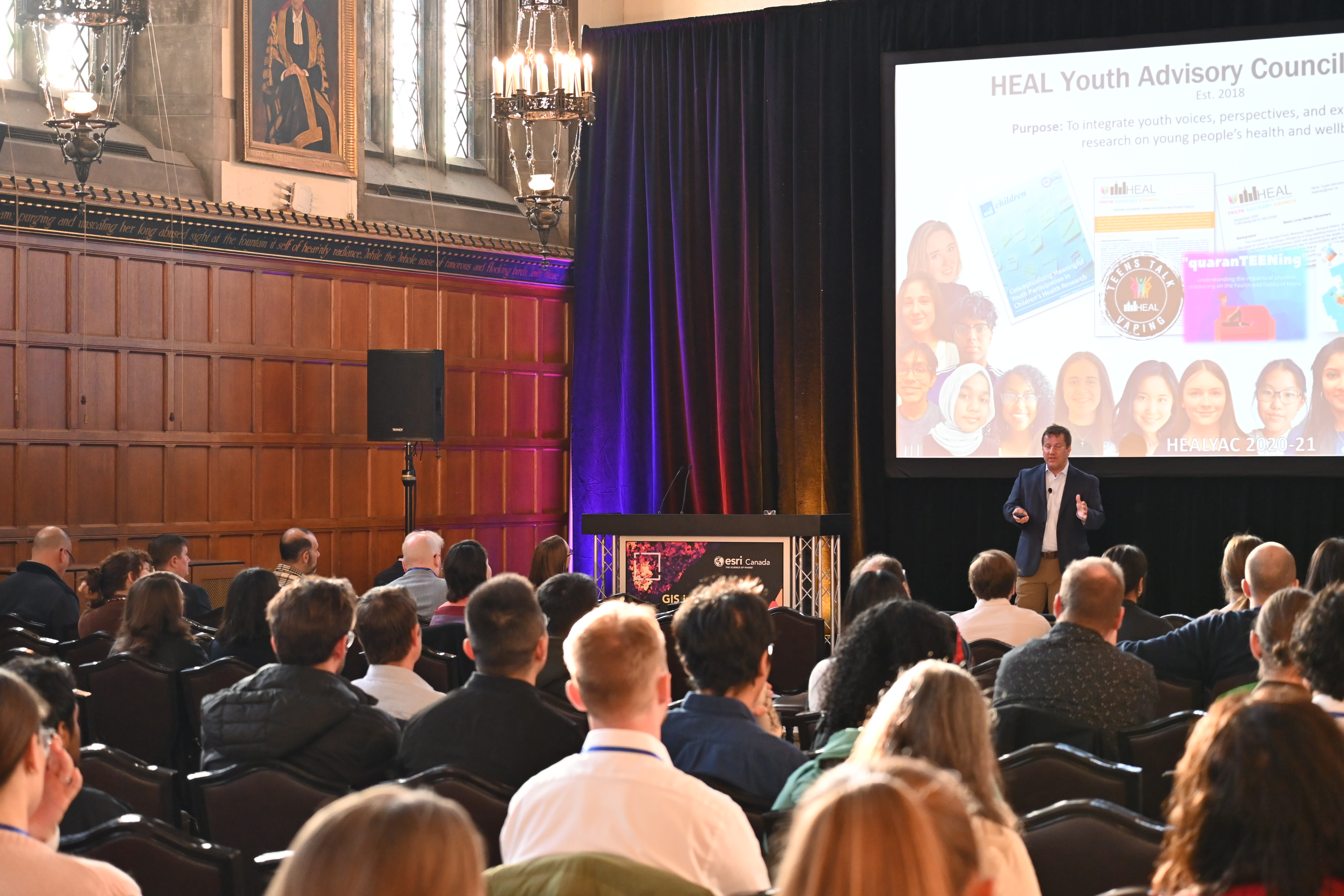 A man standing on stage, in front of a screen speaking to a large group of people.