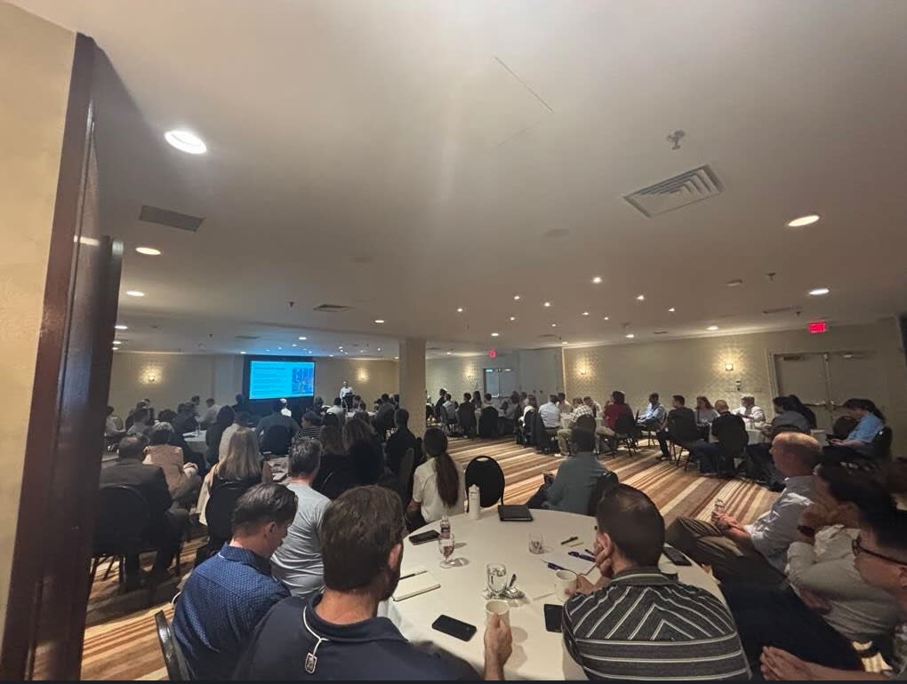 A meeting room with a dozen or more round tables, each packed with seated professionals, arranged around the perimeter. A screen at the far end of the room shows presentation slides. The 50+ attendees are all focused on the slides and the person presenting them.