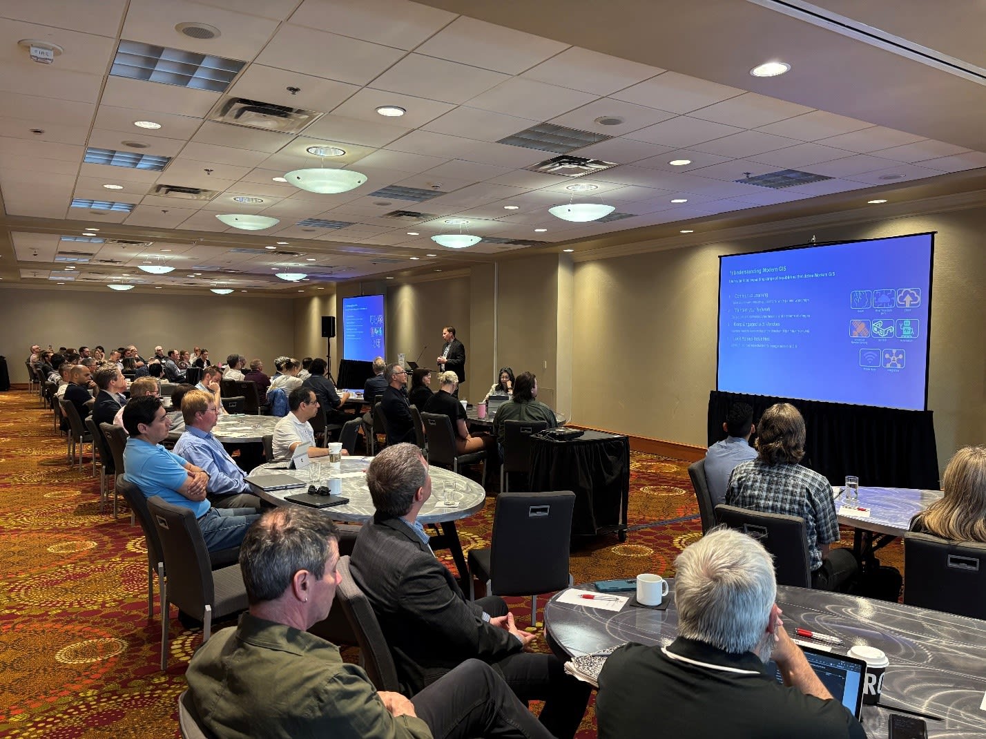 A meeting room with a number of round tables, each packed with GIS professionals, arranged in two long rows. A speaker stands before the gathered people, discussing a slide that appears on two screens, one to the left of him and one to the right. The several dozen attendees are focused on the slides.