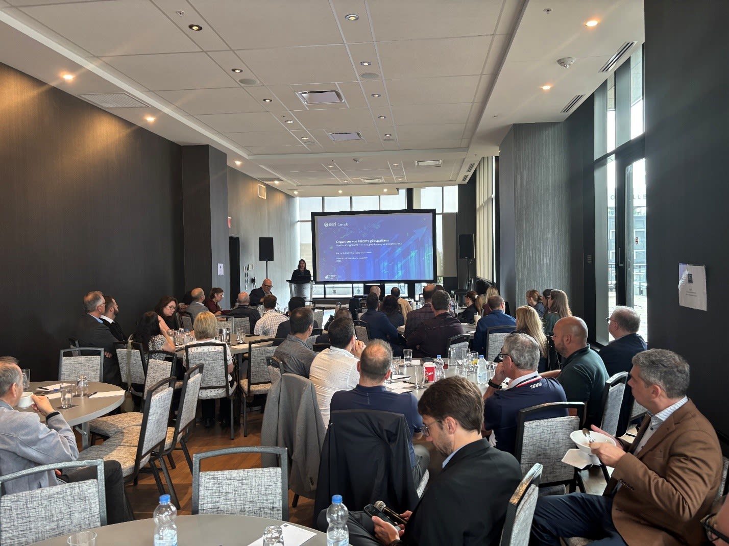 A brightly lit meeting room with floor-to-ceiling windows. Well-dressed professionals sit at round tables watching the presenter at the front of the room, taking notes and enjoying mid-morning snacks. The speaker at the podium at the front of the room addresses the crowd while a blue presentation slide is displayed on the large screen next to her.