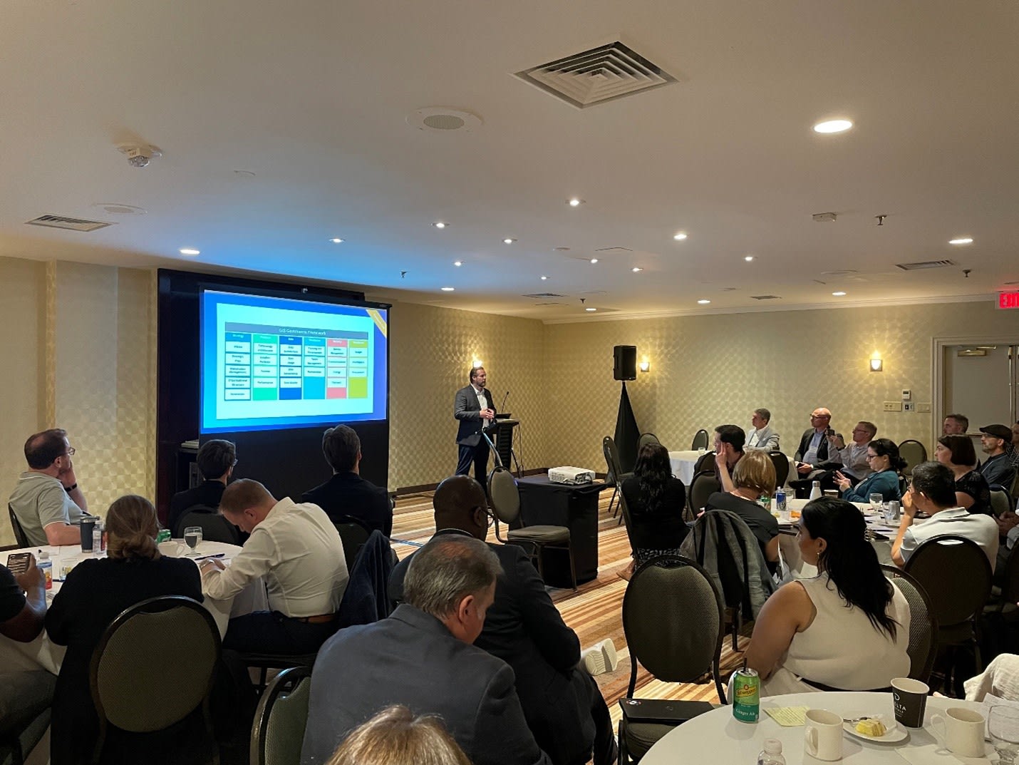 A meeting room with a number of round tables, each packed with seated professionals, arranged in the foreground. A speaker stands at a podium at the front of the room, discussing a slide that is currently displayed on the screen next to him. Some attendees are looking at the speaker while others are taking notes intently.