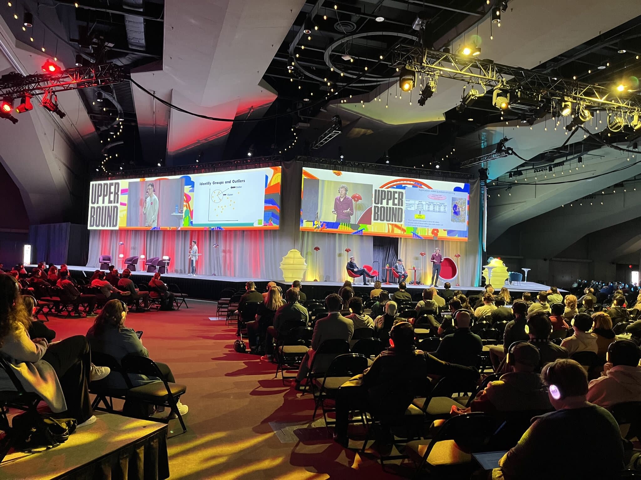 Photo prise à l’arrière d’une grande salle, montrant des rangées de participants assis regardant la scène à l’avant, où se trouvent les présentateurs ainsi que de grands écrans affichant les intervenants et le contenu de la présentation.