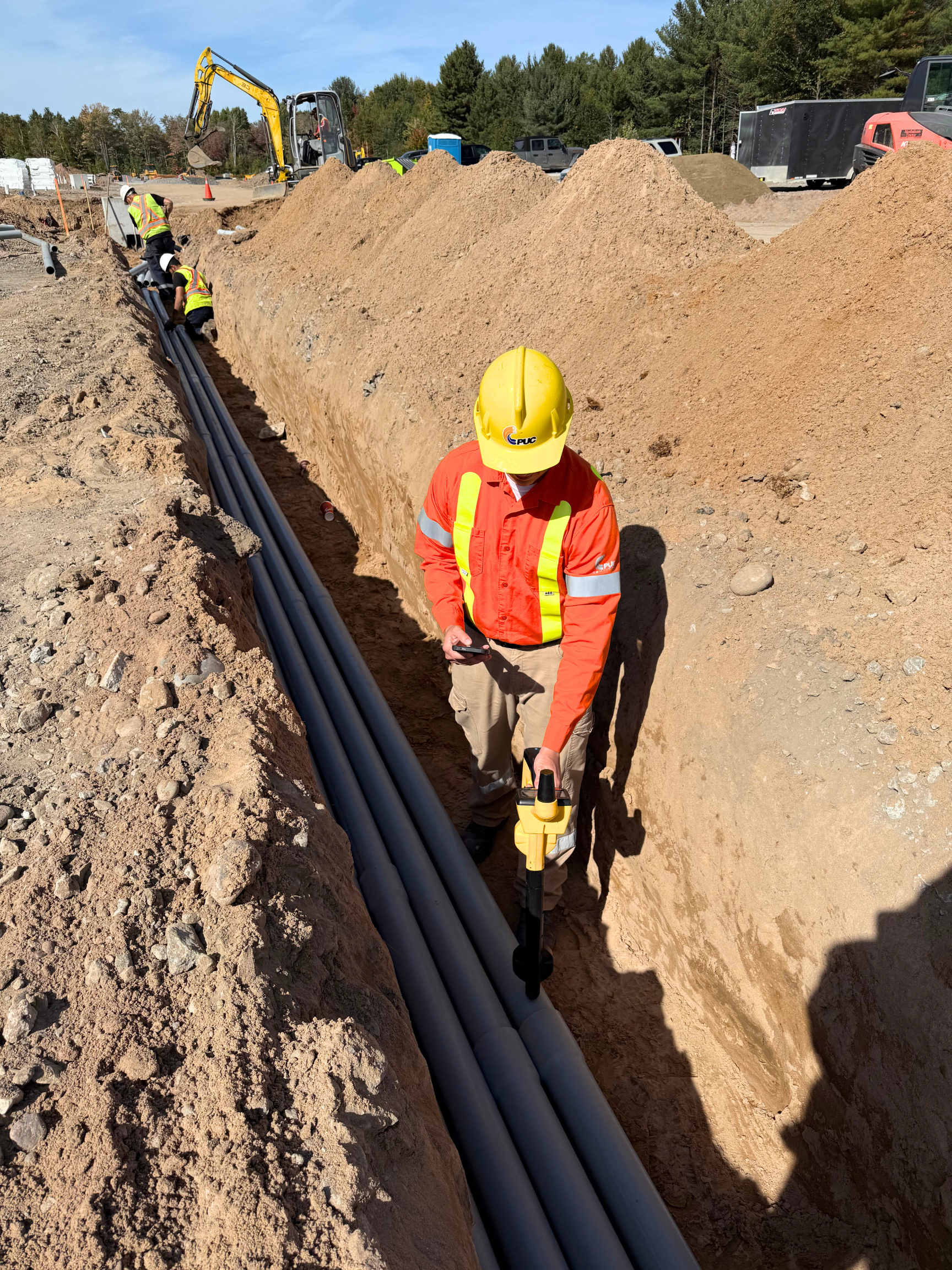 PUC locator wearing reflective gear and a hard hat maps underground infrastructure with a locate device in a long ditch on a sunny day.