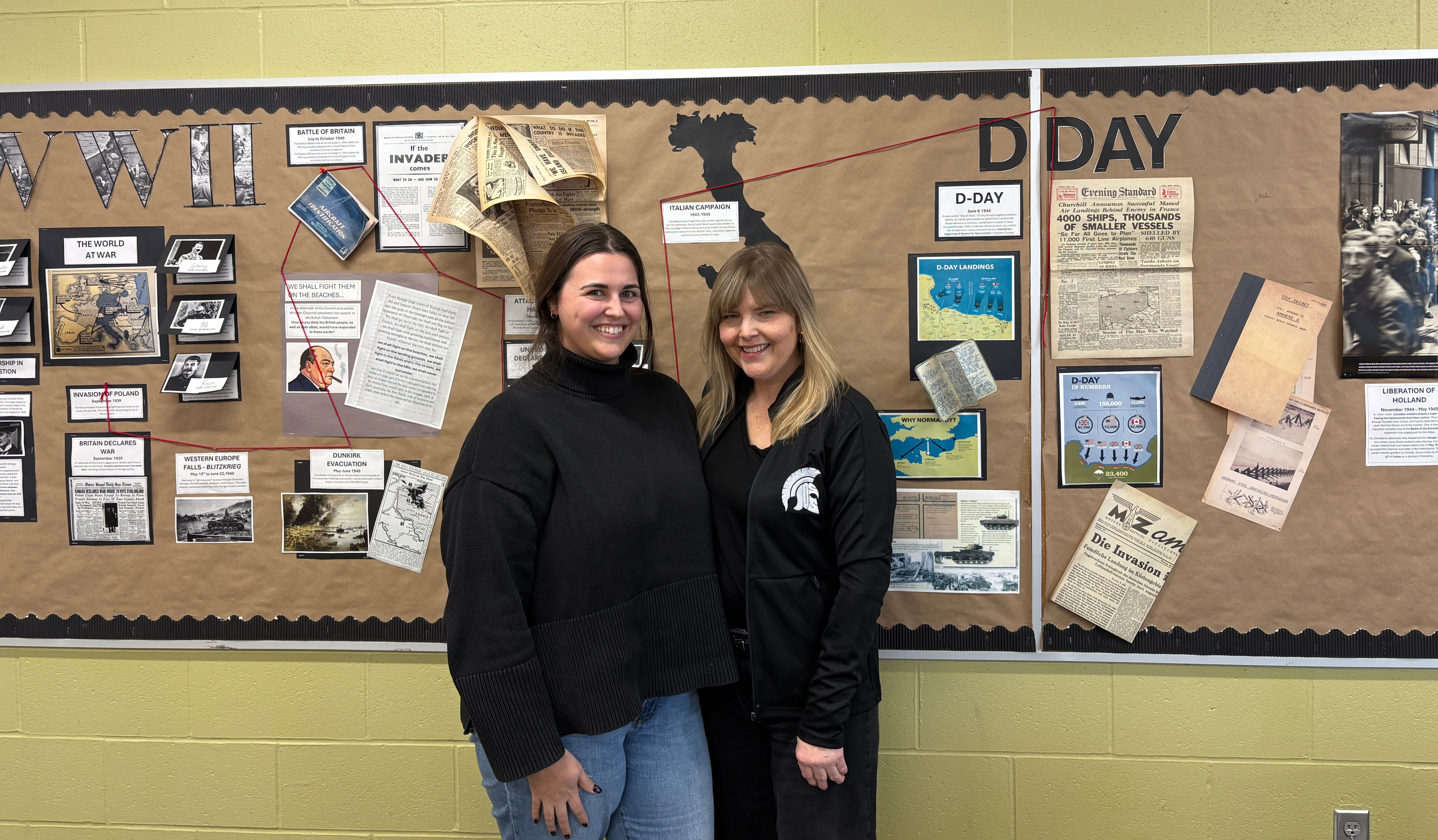 Deux femmes souriant devant un tableau d&rsquo;affichage avec des travaux d&rsquo;&eacute;l&egrave;ves portant sur l&rsquo;histoire du Jour&nbsp;J.