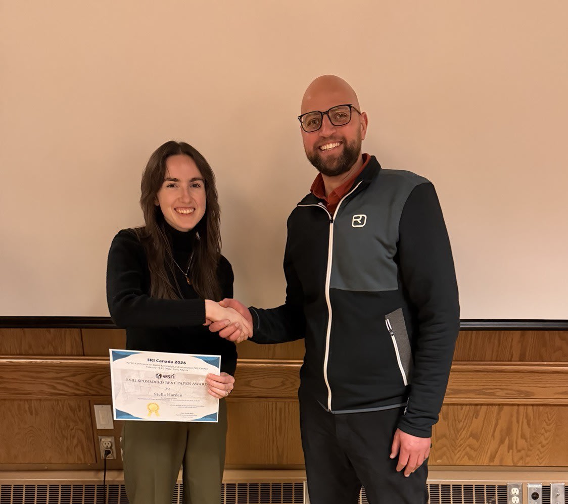 Stella Harden stands smiling and shaking hands with the award presenter, Mohamed Ahmed from Esri Canada, while holding her Best Paper Award certificate at the SKI Canada 2026 conference in Banff, Alberta.