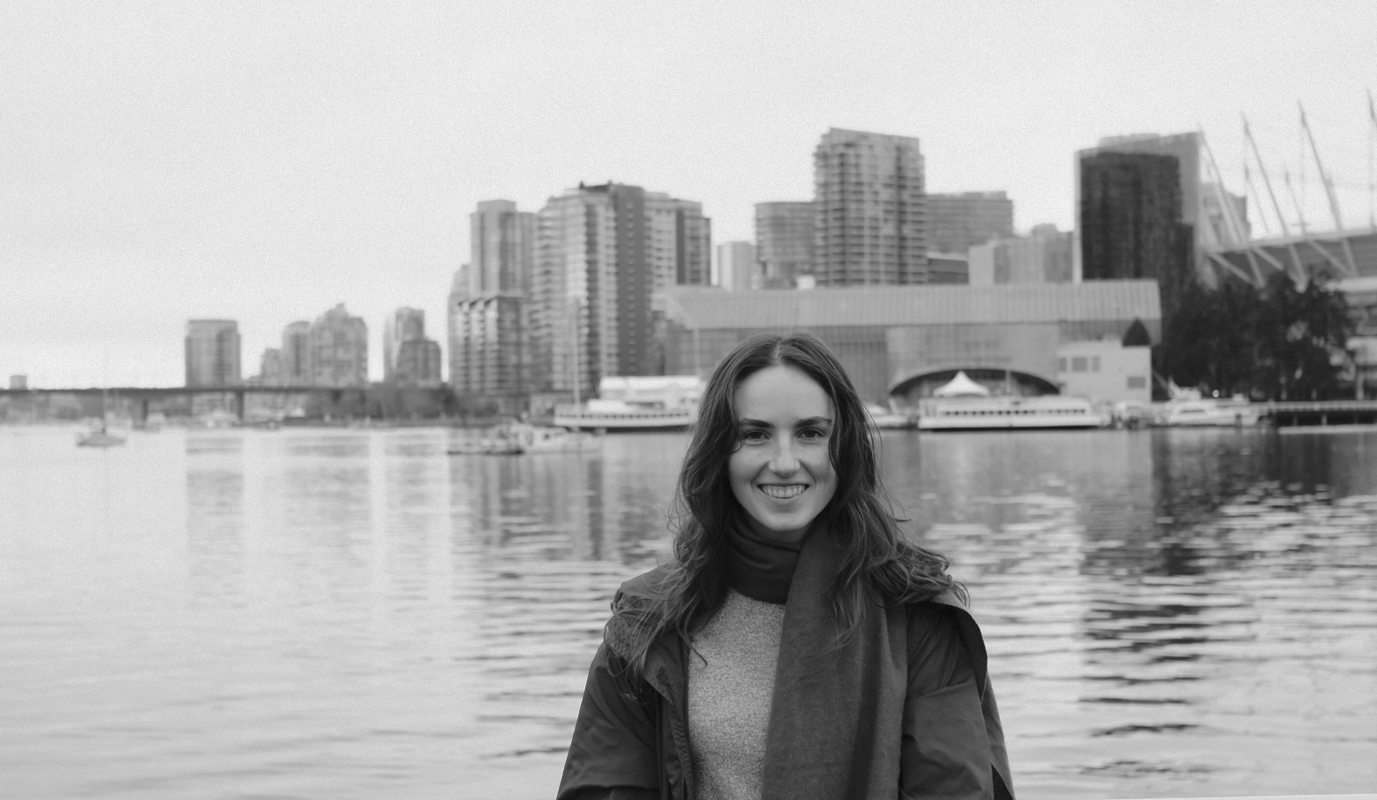 Black-and-white photo of Stella smiling by the waterfront, wearing a coat and scarf, with boats, calm water, and Vancouver&rsquo;s downtown skyline in the background.