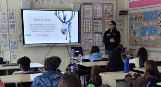A woman standing at the front of a classroom presenting information from a screen to a group of students.