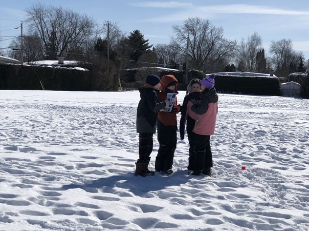 Four students in winter gear standing in field of snow huddled together, engaged in conversation.