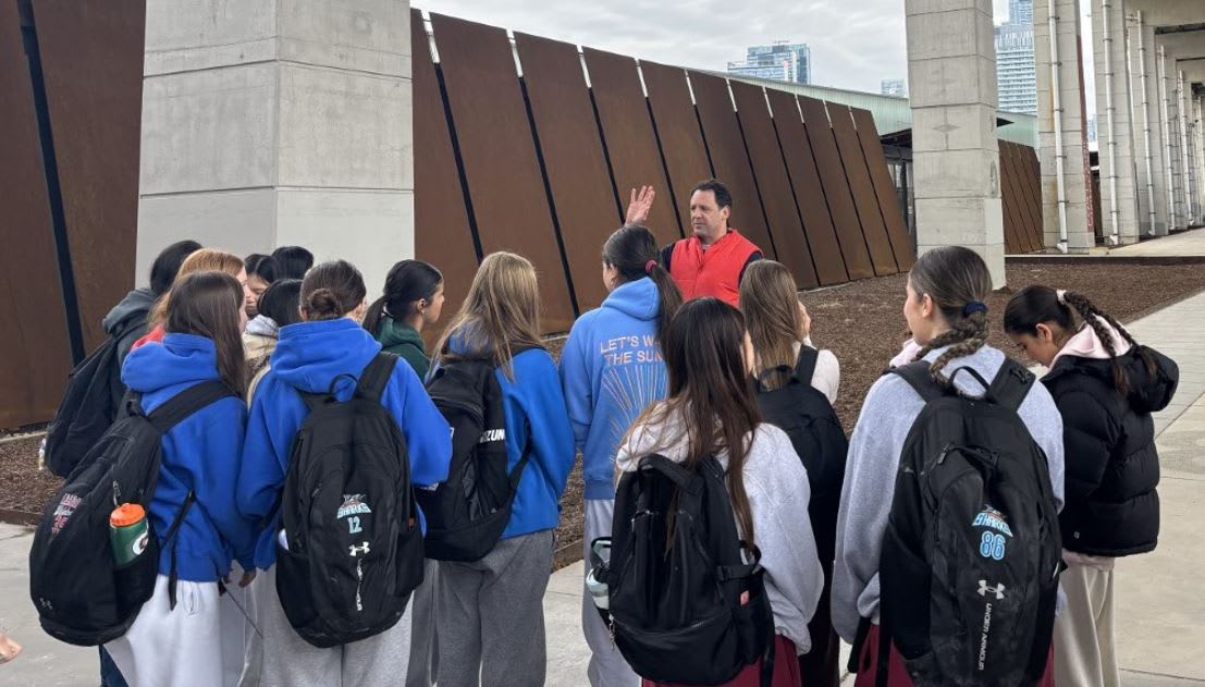 An adult male speaking to a group of a dozen female students in an urban space.