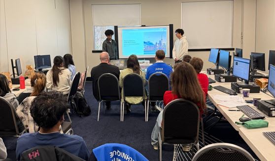 Two male high school students standing in front of a large projector screen presenting their project to about a dozen people.
